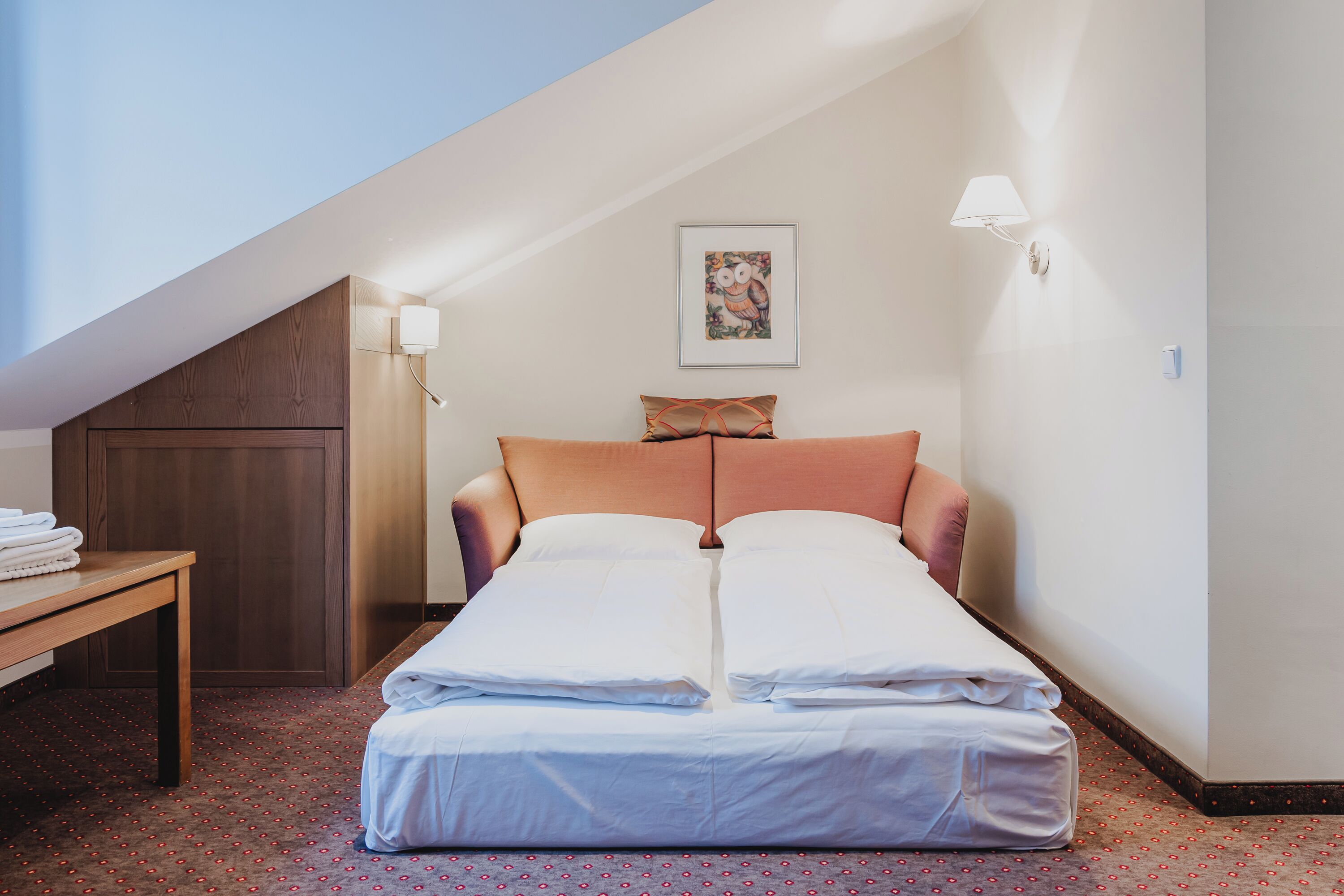 A single bed in a room with a sloped ceiling, a wooden table, and a wall-mounted lamp.
