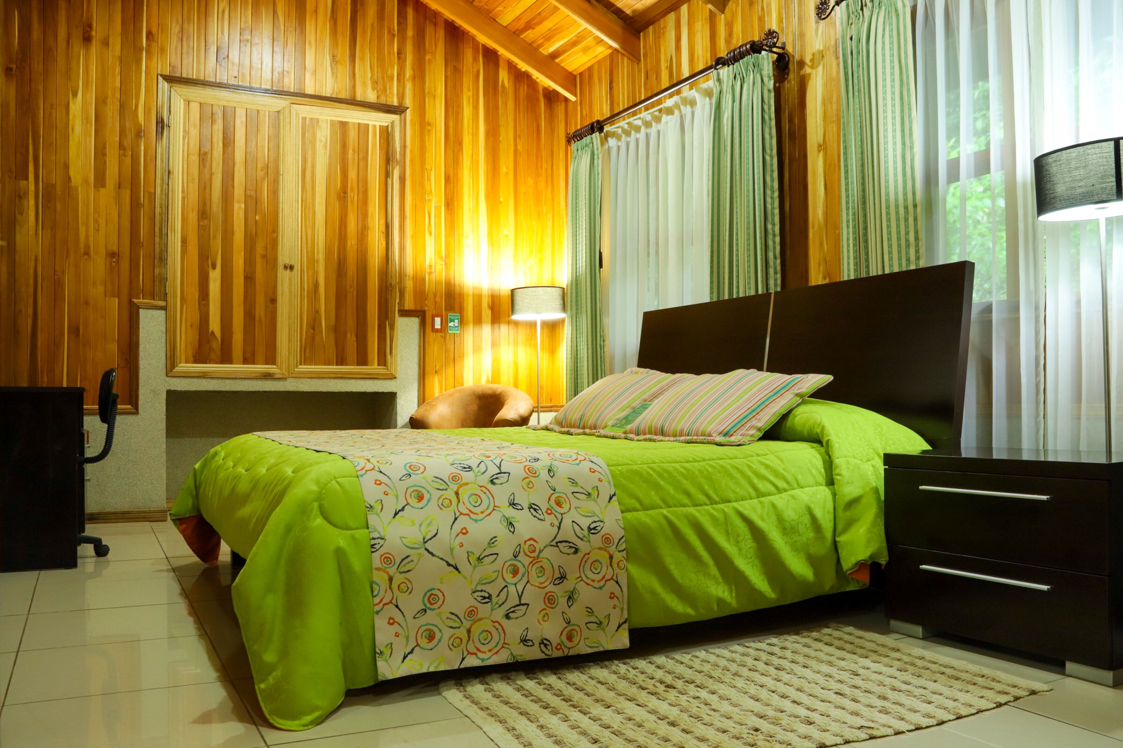 A bedroom with a wooden ceiling, a bed with a green bedspread, a nightstand, a lamp, and a window with curtains.