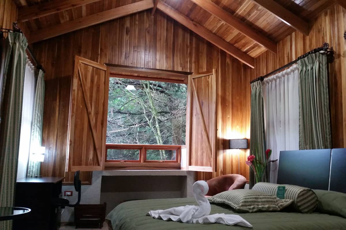 A wooden cabin bedroom with a bed, a desk, and a window overlooking a forest.