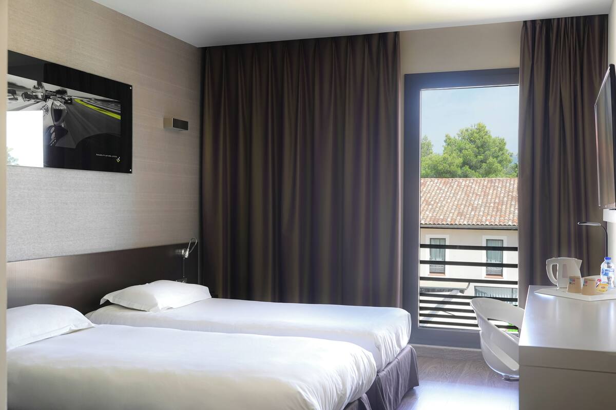 A hotel room with two beds, a desk, a chair, a large window with curtains, and a wall-mounted TV.