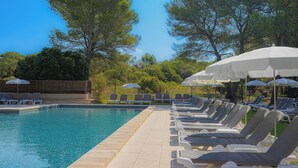 Zona de piscina con sillas de descanso y sombrillas, rodeada de árboles.