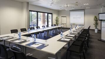 Un salón de conferencias con mesas largas, sillas, botellas de agua y una pantalla mostrando
