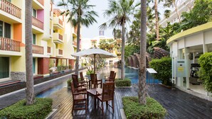 A poolside area with wooden deck seating, palm trees, and a view of a hotel building. - Mykonos Beach Apartment (Hua Hin)