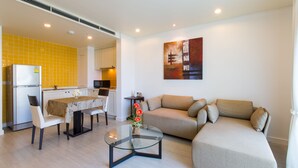 A modern living room with a glass coffee table, a beige sofa, and a dining area with a yellow-tiled backsplash and a refrigerator. - Mykonos Beach Apartment (Hua Hin)