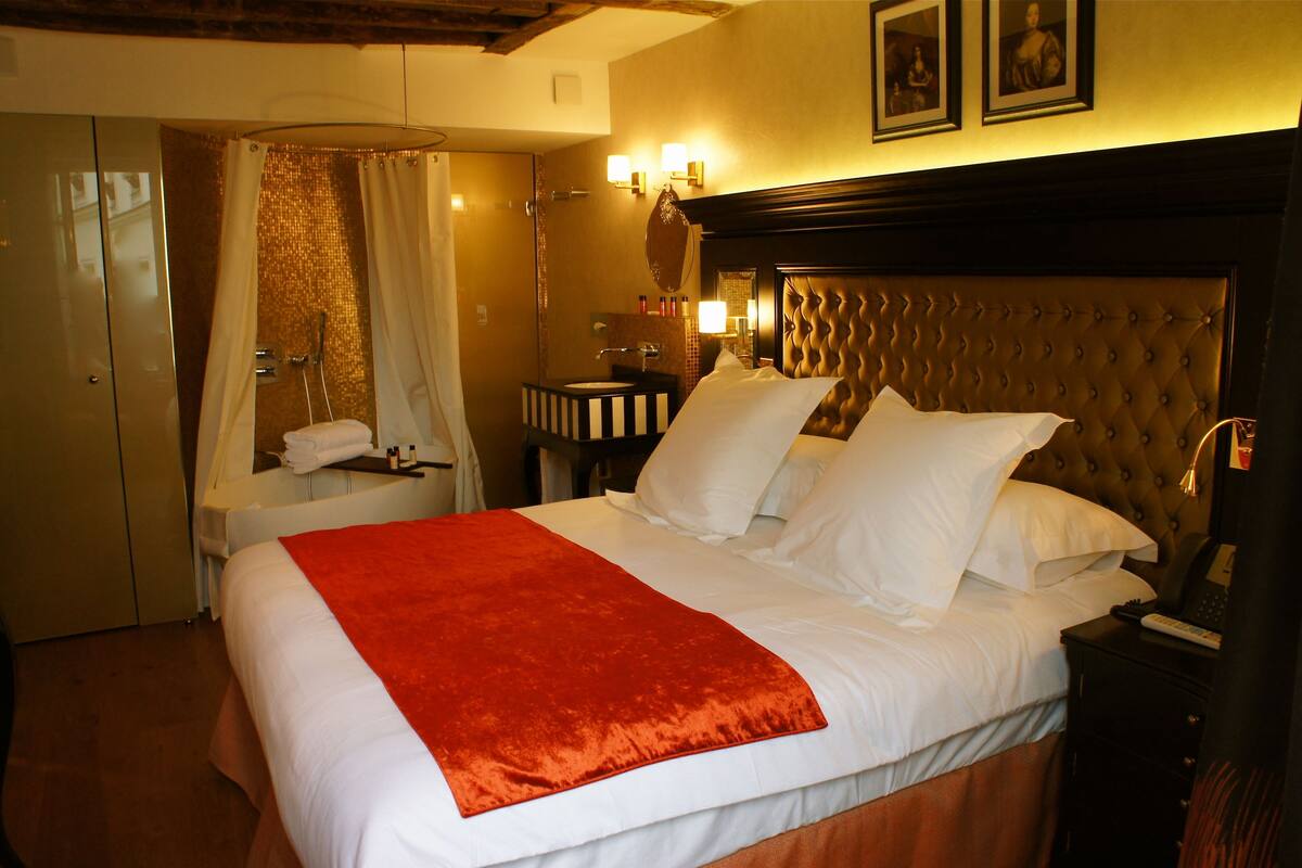 A hotel room with a large bed, a bedside table, a nightstand, a mirror, and a shower area.