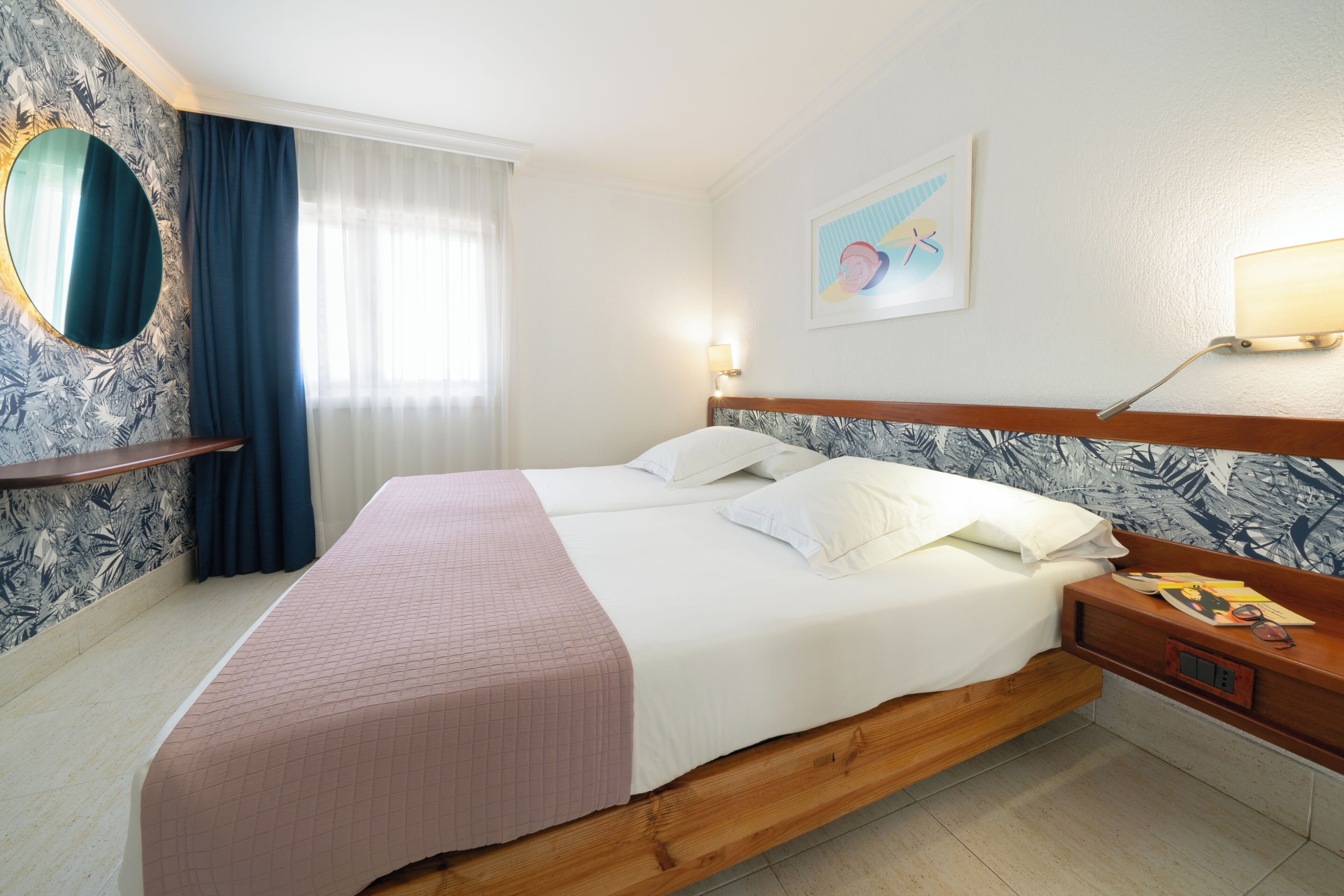 Hotelkamer met een bed, nachtkastje, spiegel, wandklok en raam met gordijnen.
