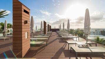 Un área de lounge en la azotea con tumbonas y sombrillas, una estructura de madera y vistas del horizonte de la ciudad.