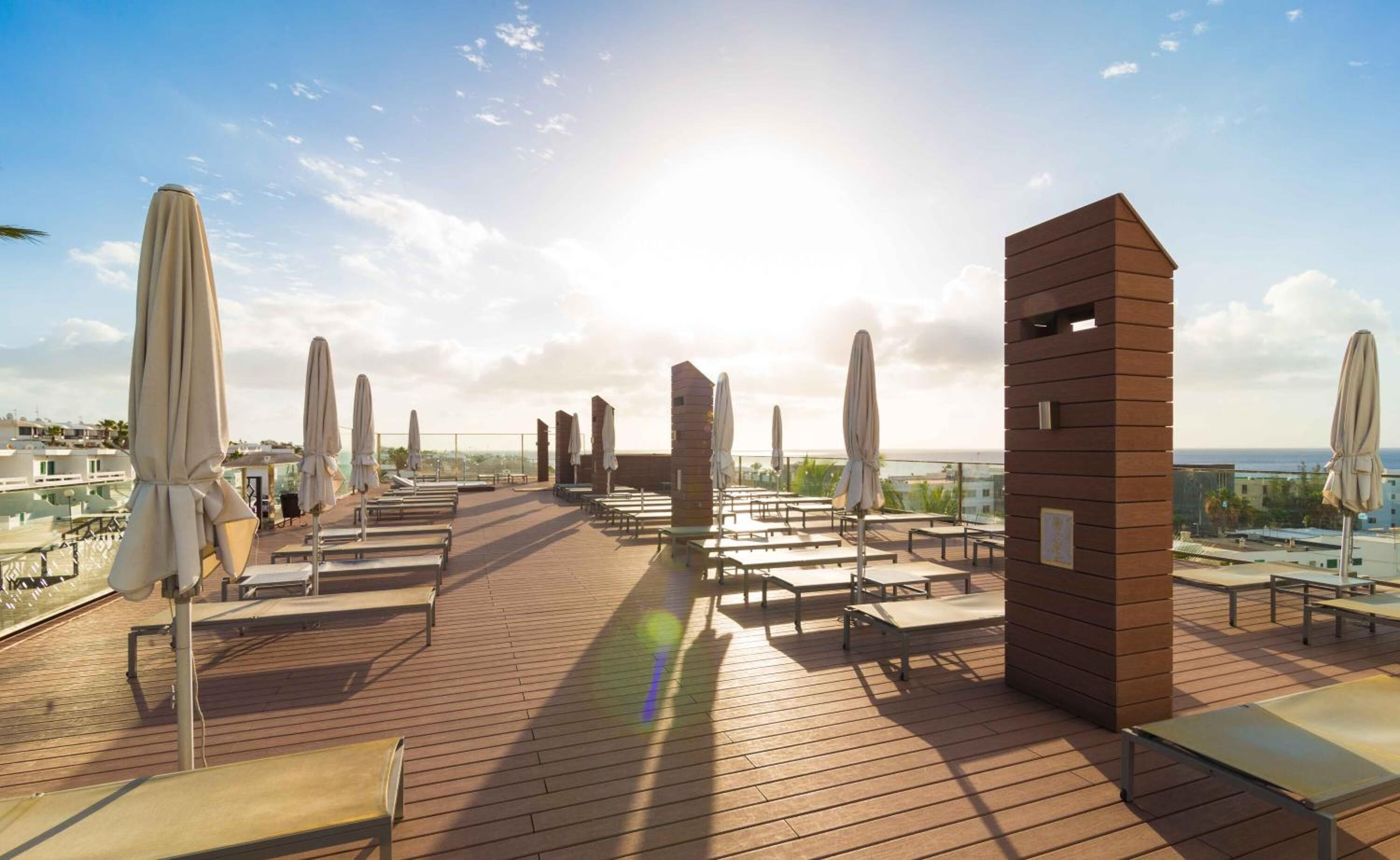 A rooftop terrace with sun loungers and umbrellas, overlooking a cityscape and the sea.
