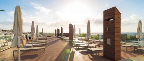 A rooftop terrace with sun loungers and umbrellas, overlooking a cityscape and the sea.