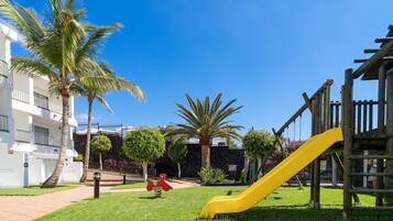Guests can enjoy a playground with a slide and climbing structures amidst a lush lawn and palm trees.