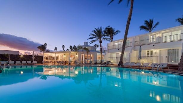 Una piscina con sillones de descanso y palmeras, rodeada por un edificio con varios balcones y ventanas amplias.