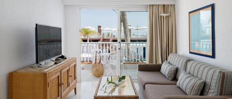 A modern living room with a flat-screen TV, wicker furniture, a sofa with striped cushions, and a view of a balcony with umbrellas.