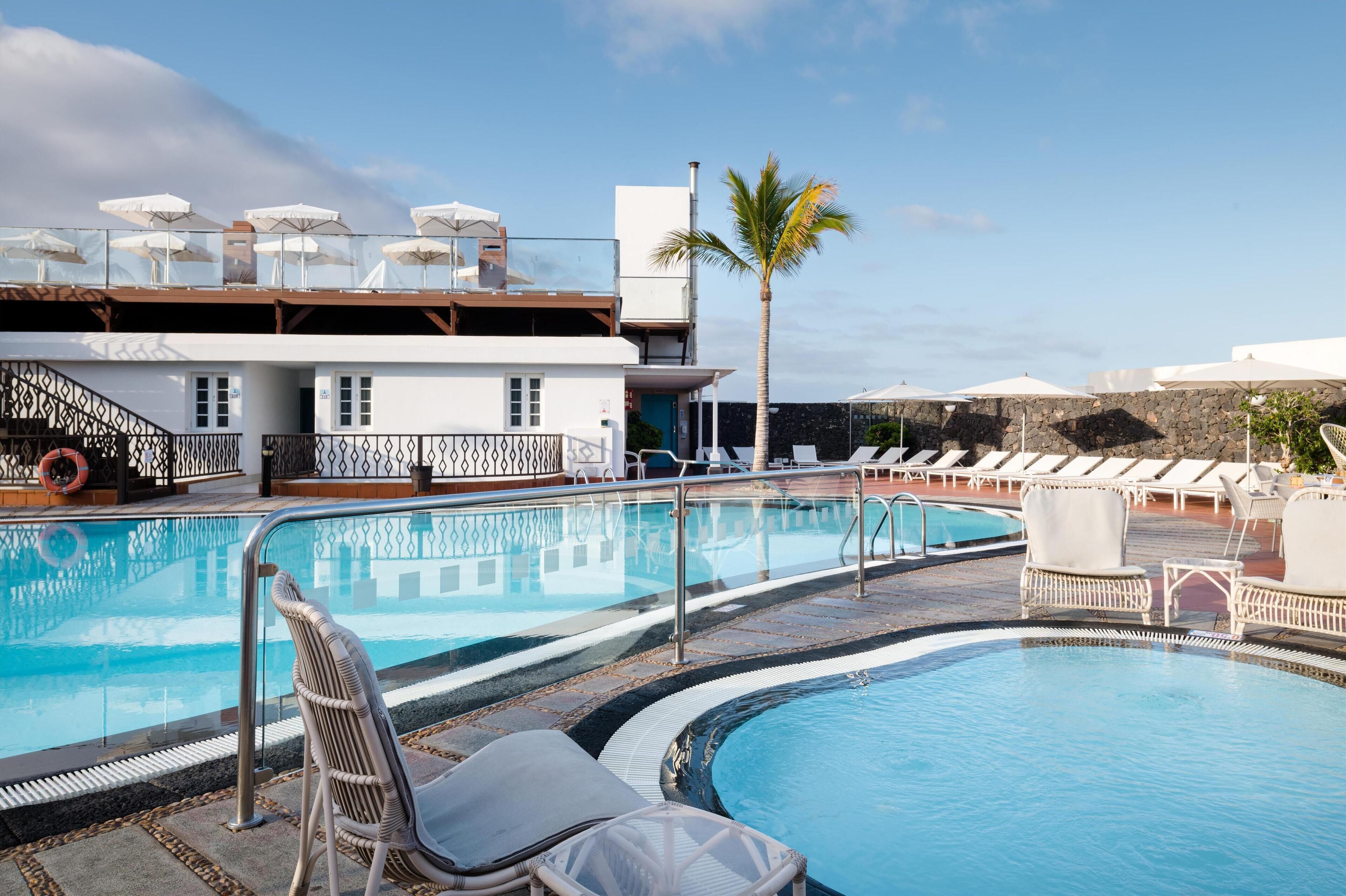 A pool area with lounge chairs and umbrellas, a palm tree, and a building with a balcony.