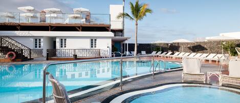 A pool area with lounge chairs and umbrellas, a palm tree, and a building with a balcony.