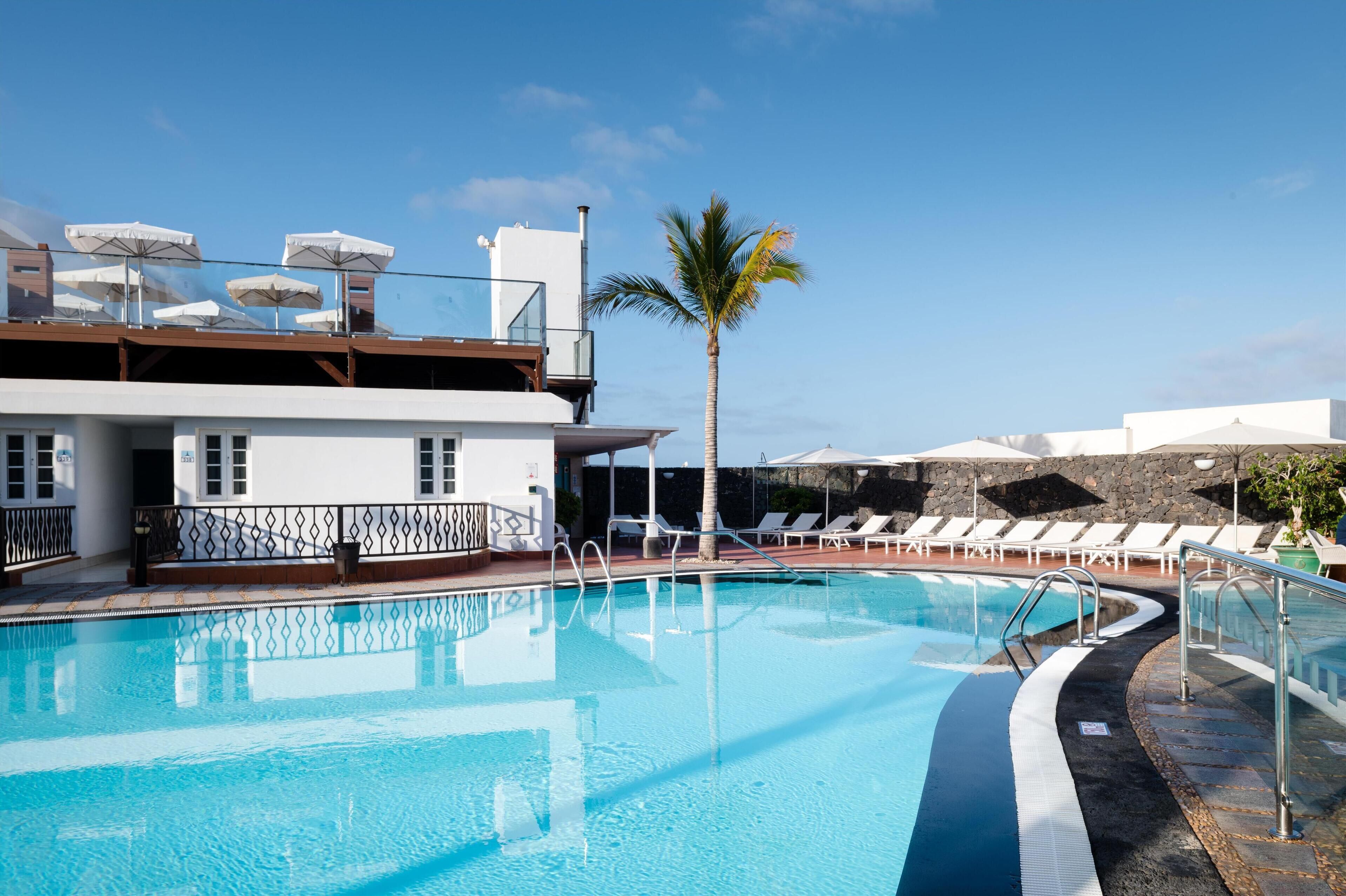 Una piscina con sillas de descanso y sombrillas, una palmera y un edificio con balcón.