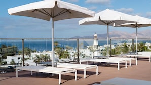 Terraza en la azotea con sillones blancos y sombrillas, con vistas a un pueblo costero y montañas.