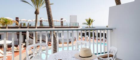 Een balkon met witte stoelen, een tafel en een hoed. Achtergrond toont een zwembad met ligbedden en parasols.
