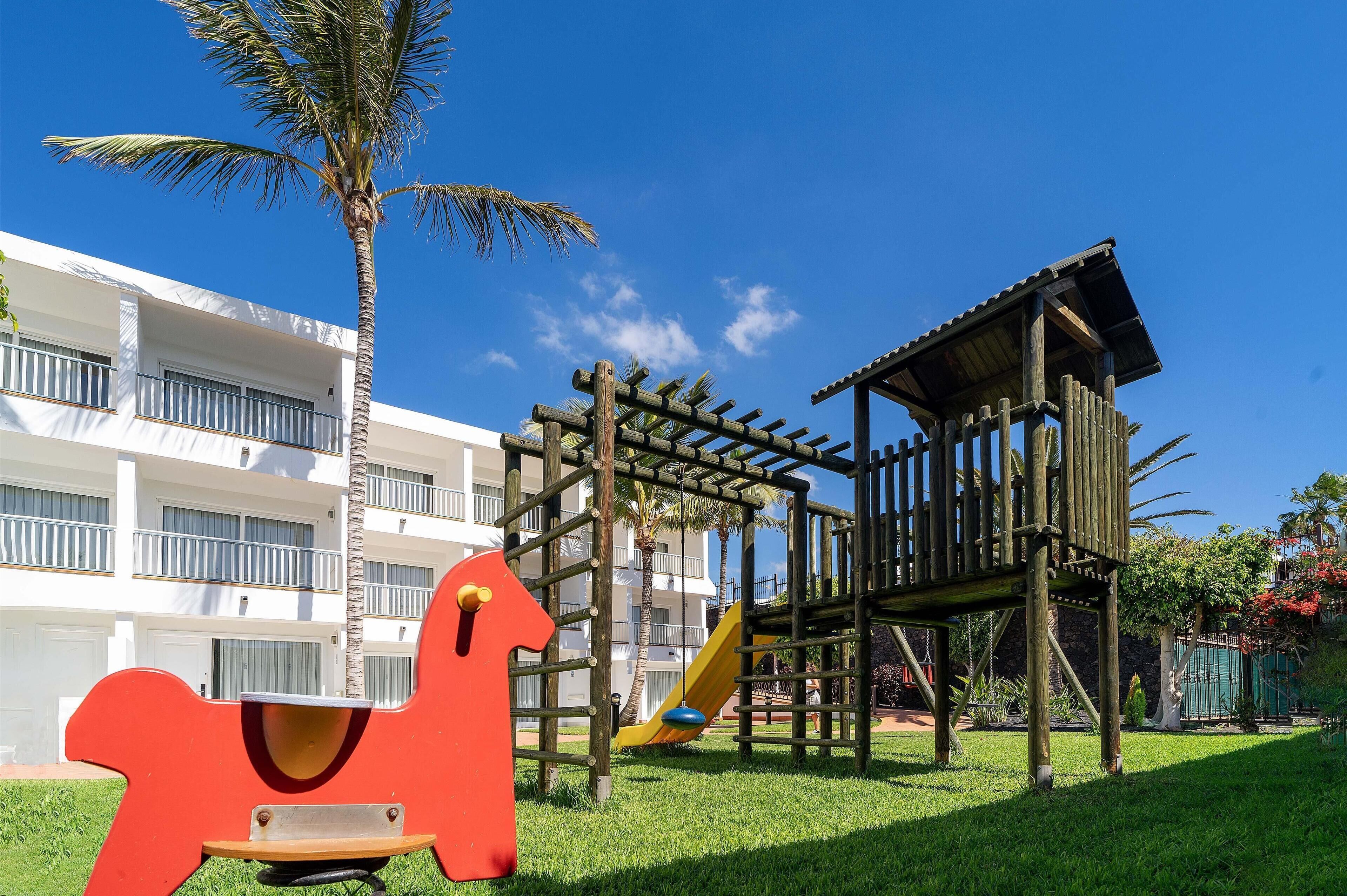 A playground with a slide and climbing structures, surrounded by a white building with balconies.