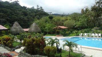 A pool area with a stone border, thatched-roof structures, and white plastic chairs.