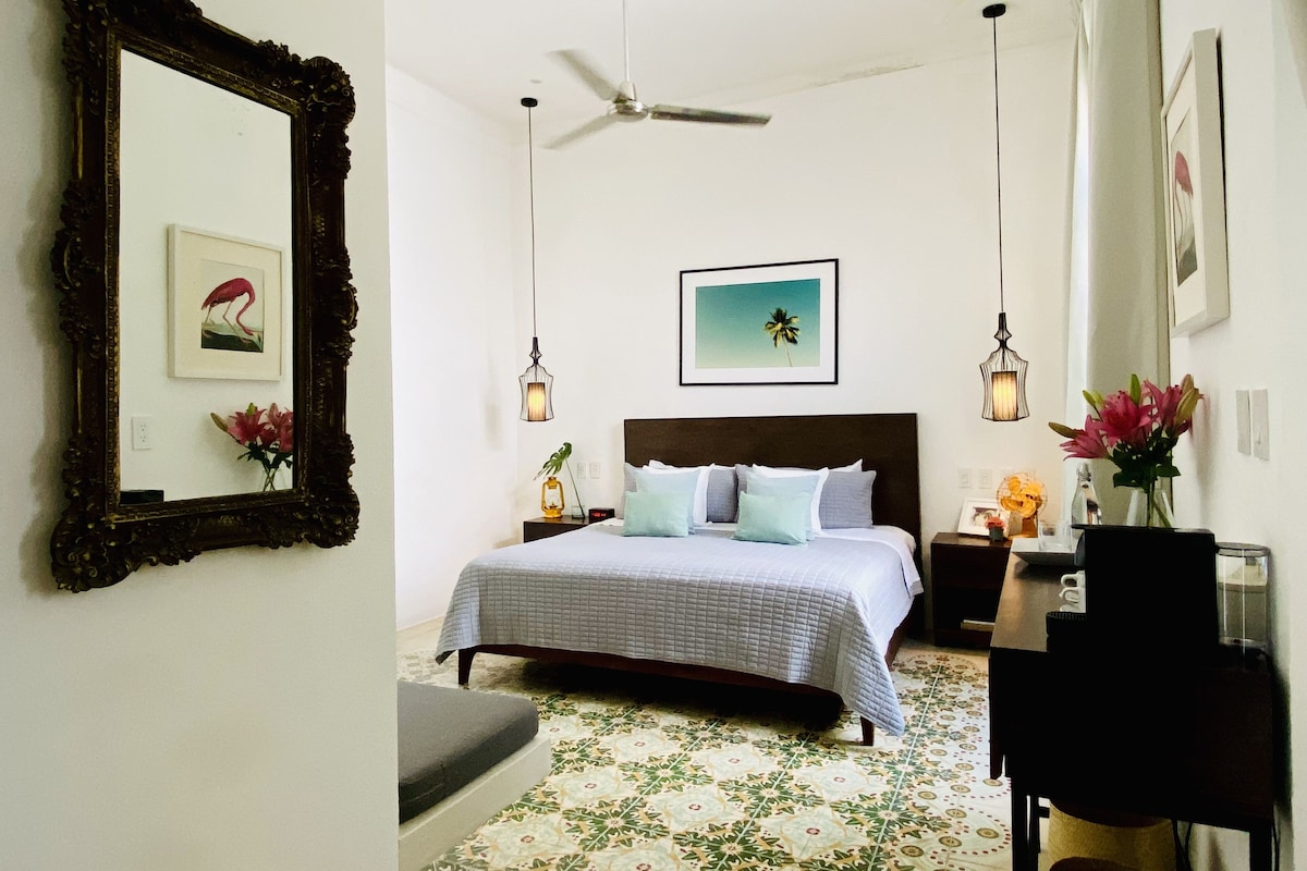 Un dormitorio con cama, ventilador de techo, escritorio con silla y espejo en la pared.
