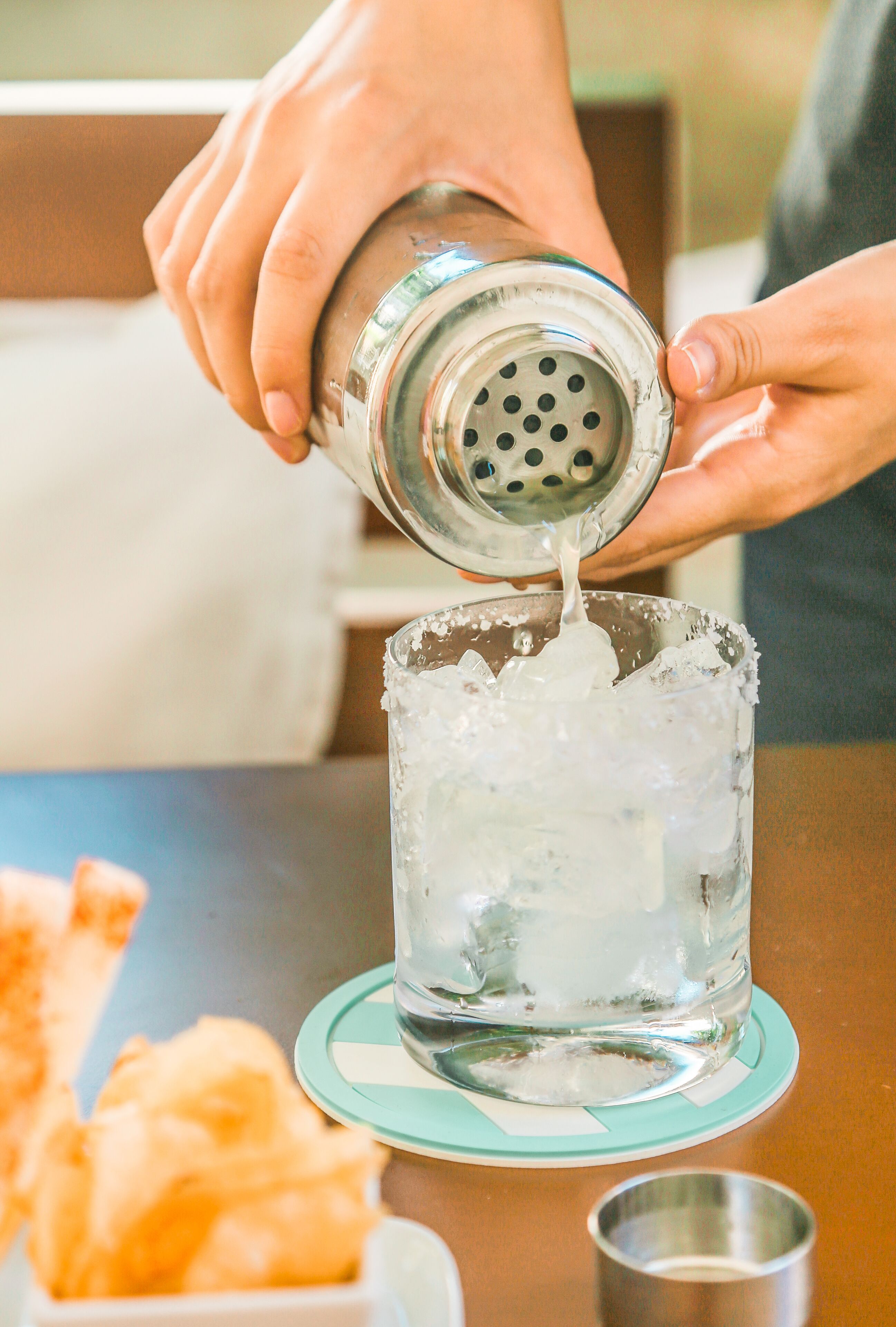 Una persona sirviendo una bebida desde un coctelera en un vaso con hielo.