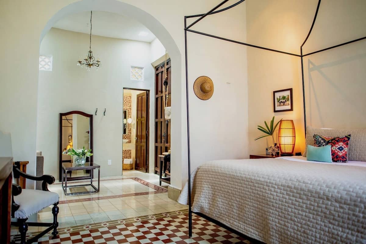 A bedroom with a four-poster bed, a bedside table with a lamp, a mirror, and a checkered floor.