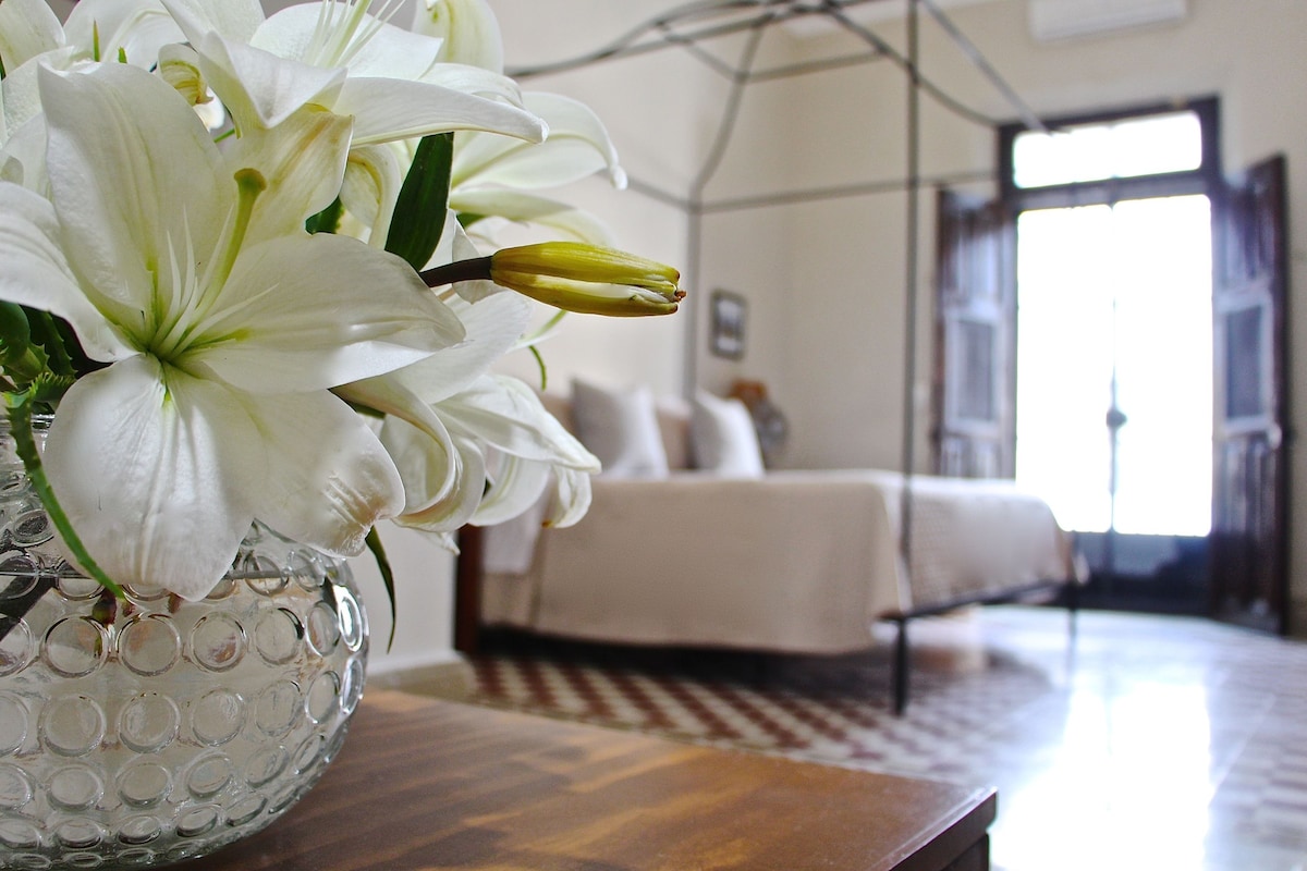 Una habitación con una cama, un florero con flores blancas y una ventana.