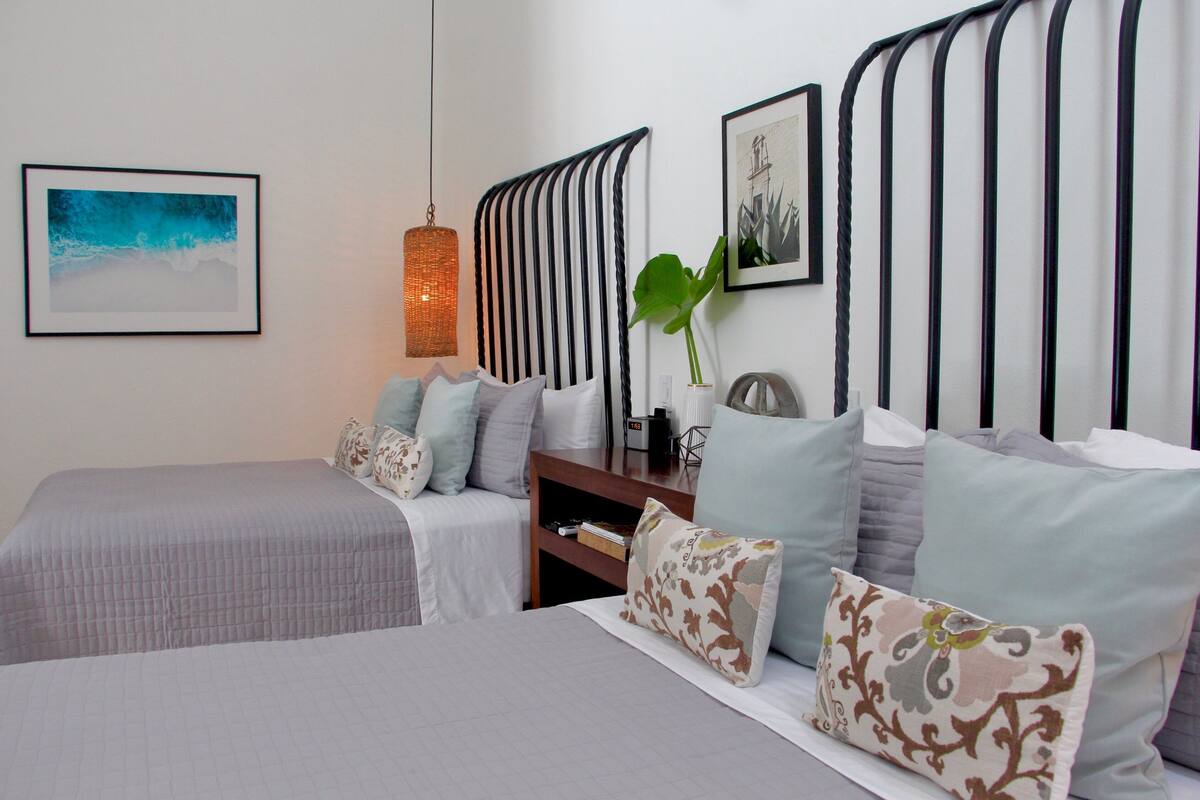 A neatly arranged bedroom with two beds, a bedside table, a hanging light fixture, and a wall-mounted artwork.