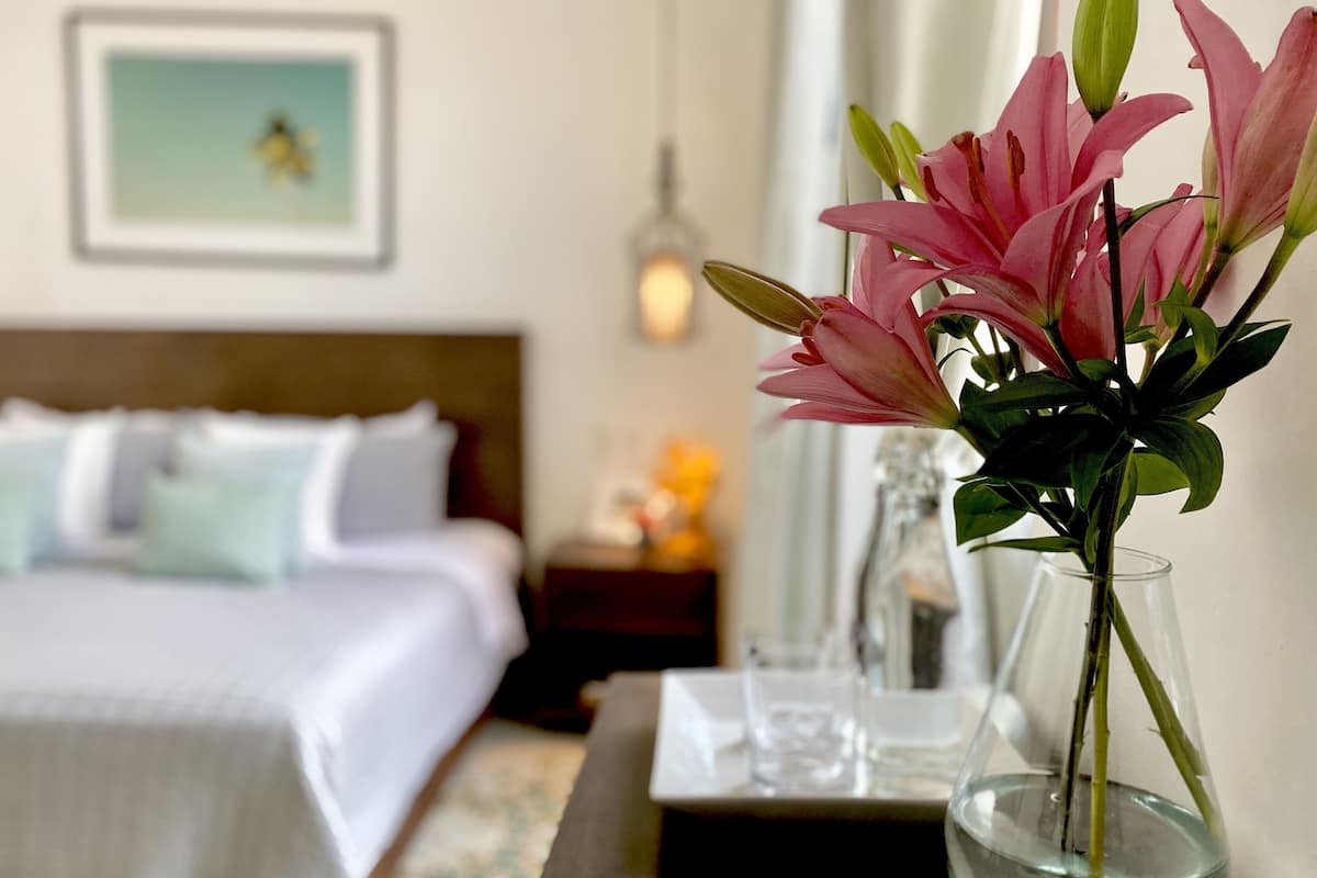 A neatly made bed with white linens, a bedside table with a vase of pink flowers, and a framed picture on the wall.
