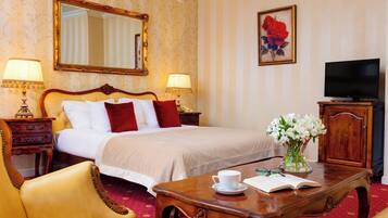 A hotel room with a large bed, two bedside lamps, a television, a wooden cabinet, a desk with a cup of coffee and an open book, a chair, and a vase of flowers.