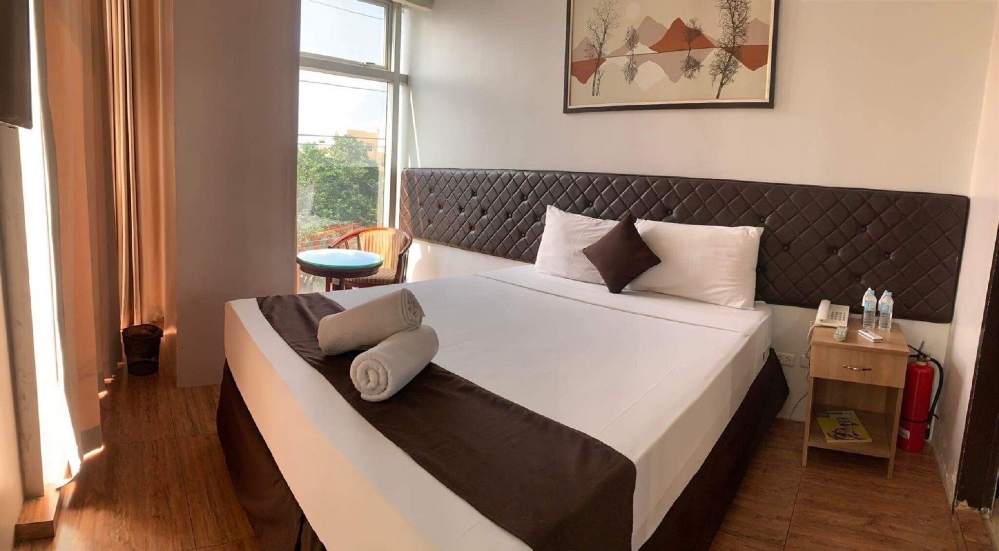 Une chambre d’hôtel avec un grand lit, une table de chevet avec un téléphone et des bouteilles d’eau, une chaise et une fenêtre donnant sur des arbres.
