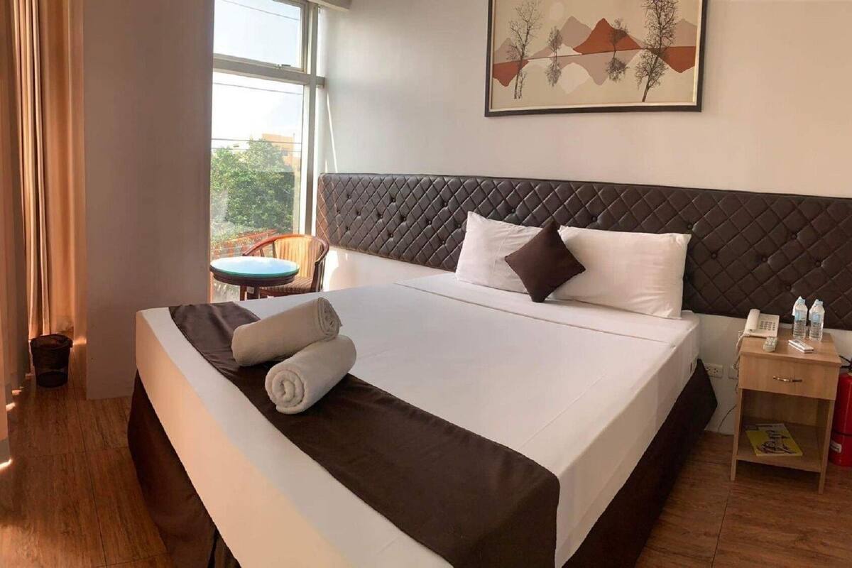 A hotel room with a large bed, a bedside table with a phone and water bottles, a chair, and a window with a view of trees.