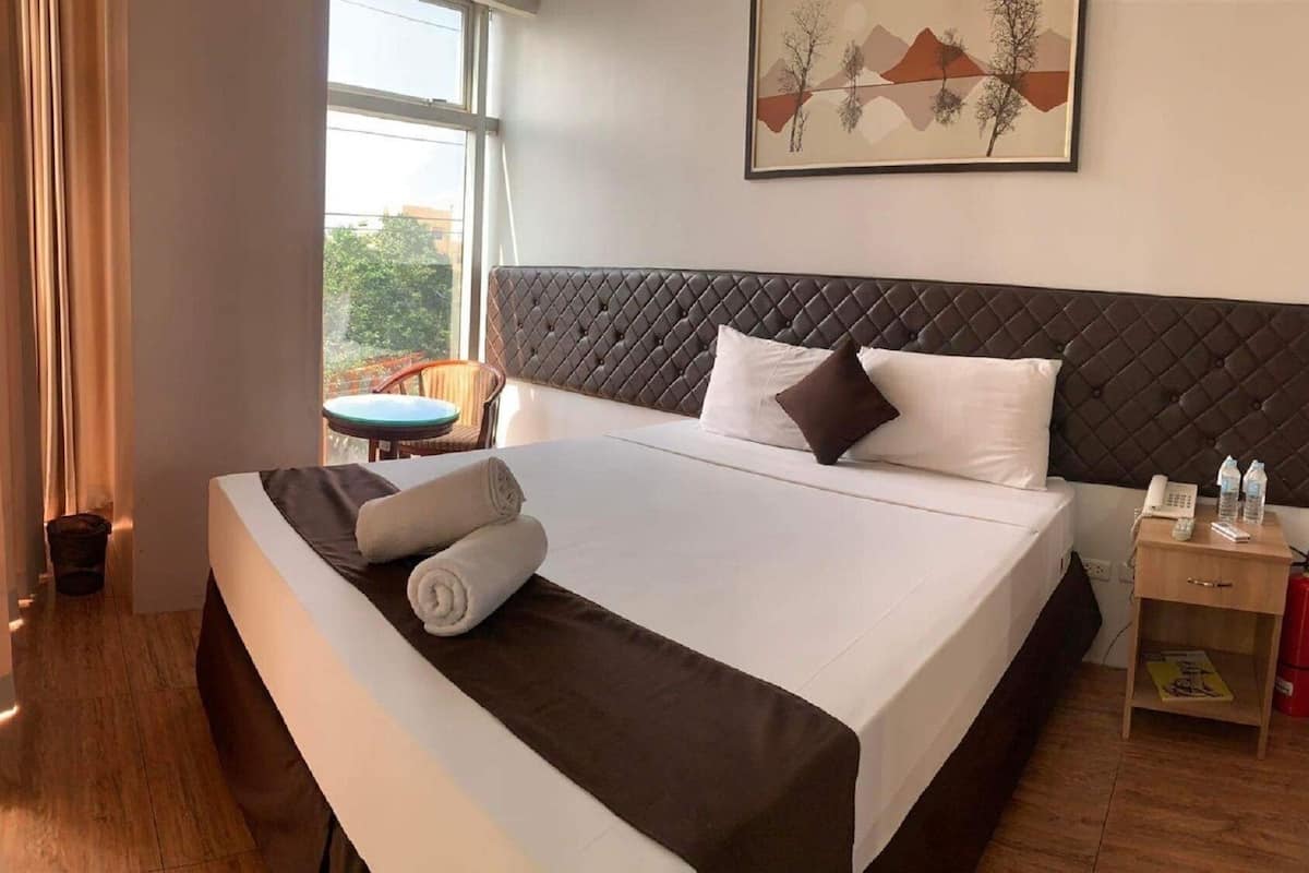 Une chambre d’hôtel avec un grand lit, une table de chevet avec un téléphone et des bouteilles d’eau, une chaise et une fenêtre donnant sur des arbres.
