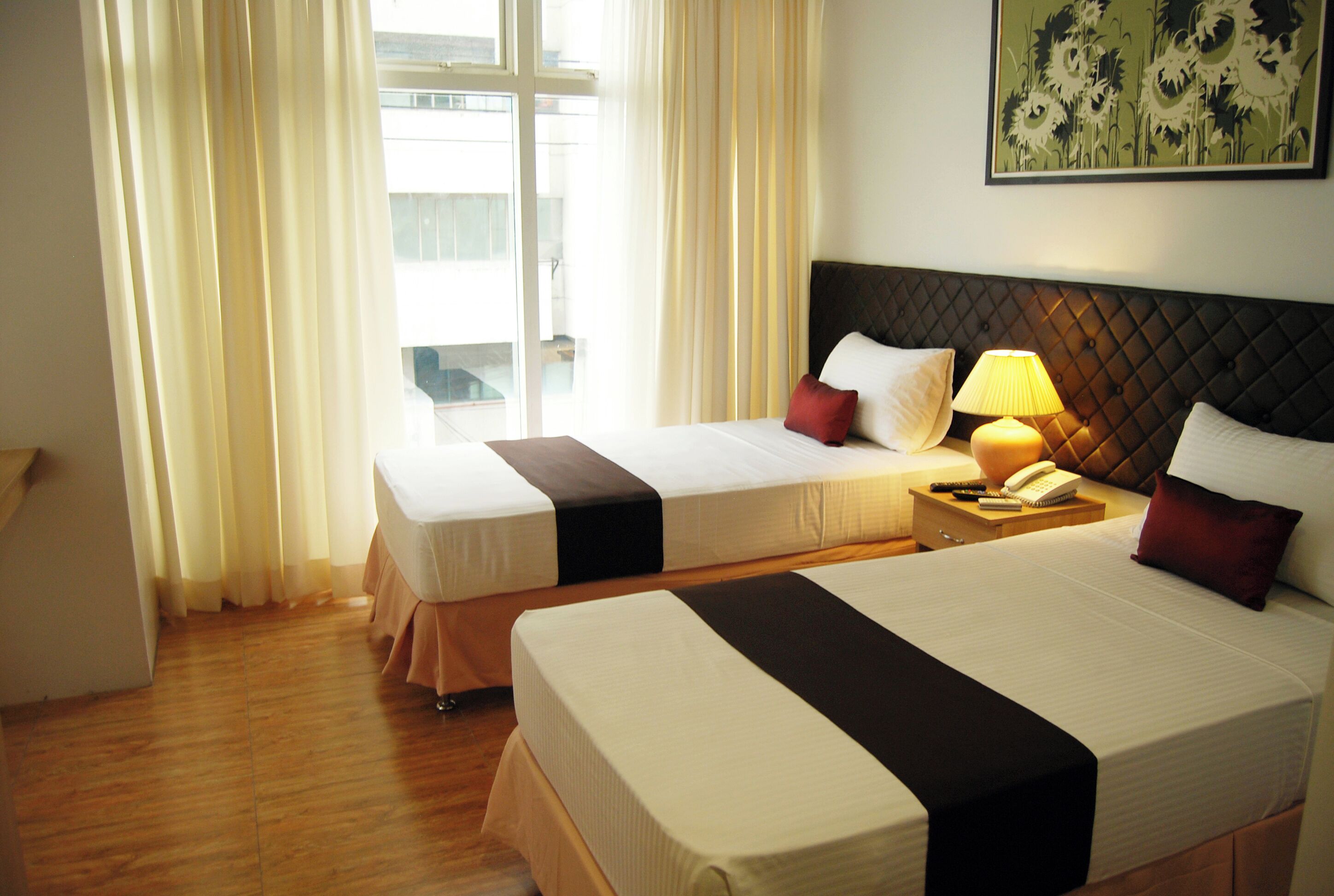 Une chambre d’hôtel avec deux lits, une fenêtre avec des rideaux, un tableau accroché au mur et une table de chevet avec une lampe.