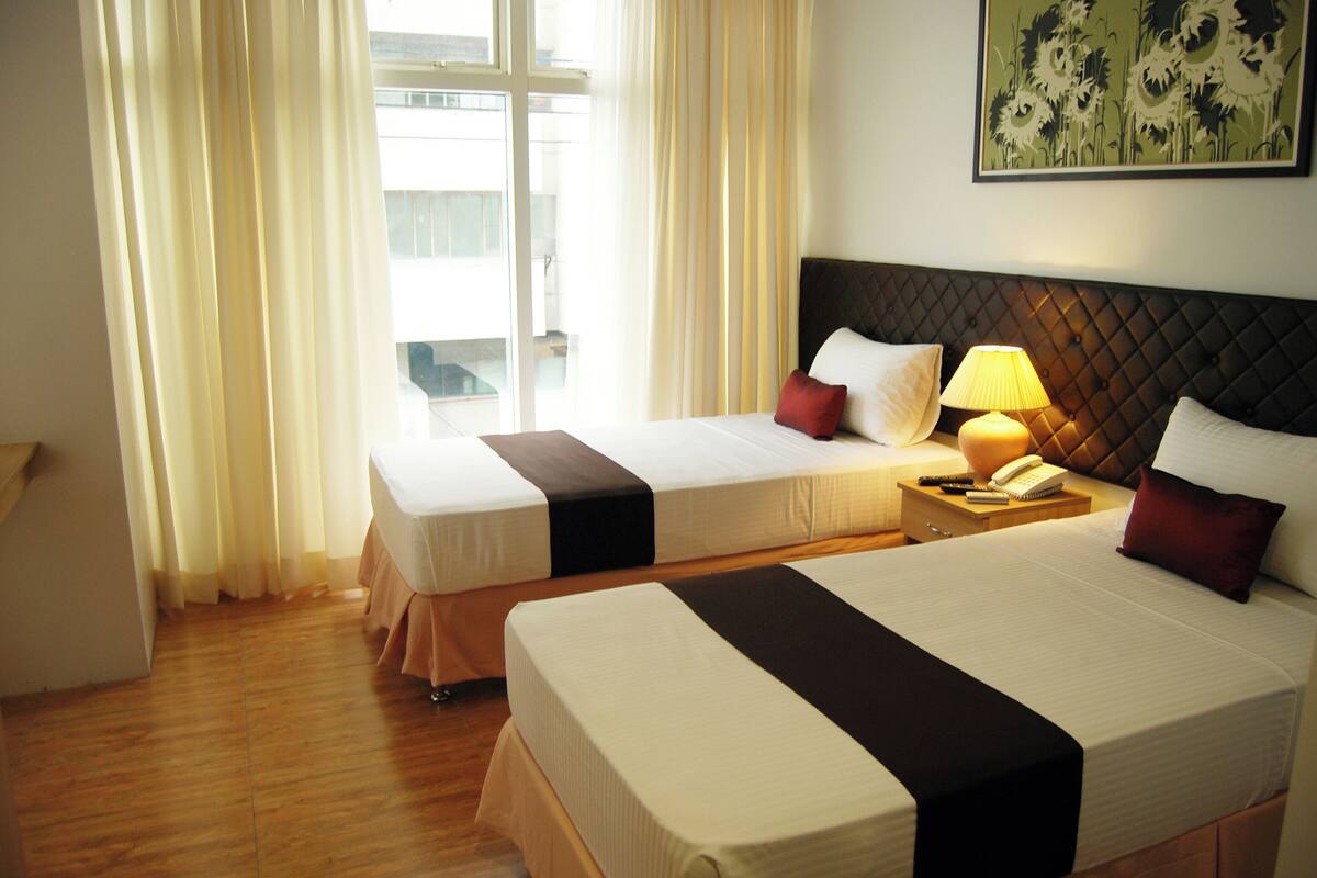 A hotel room with two beds, a window with curtains, a wall-mounted painting, and a bedside table with a lamp.