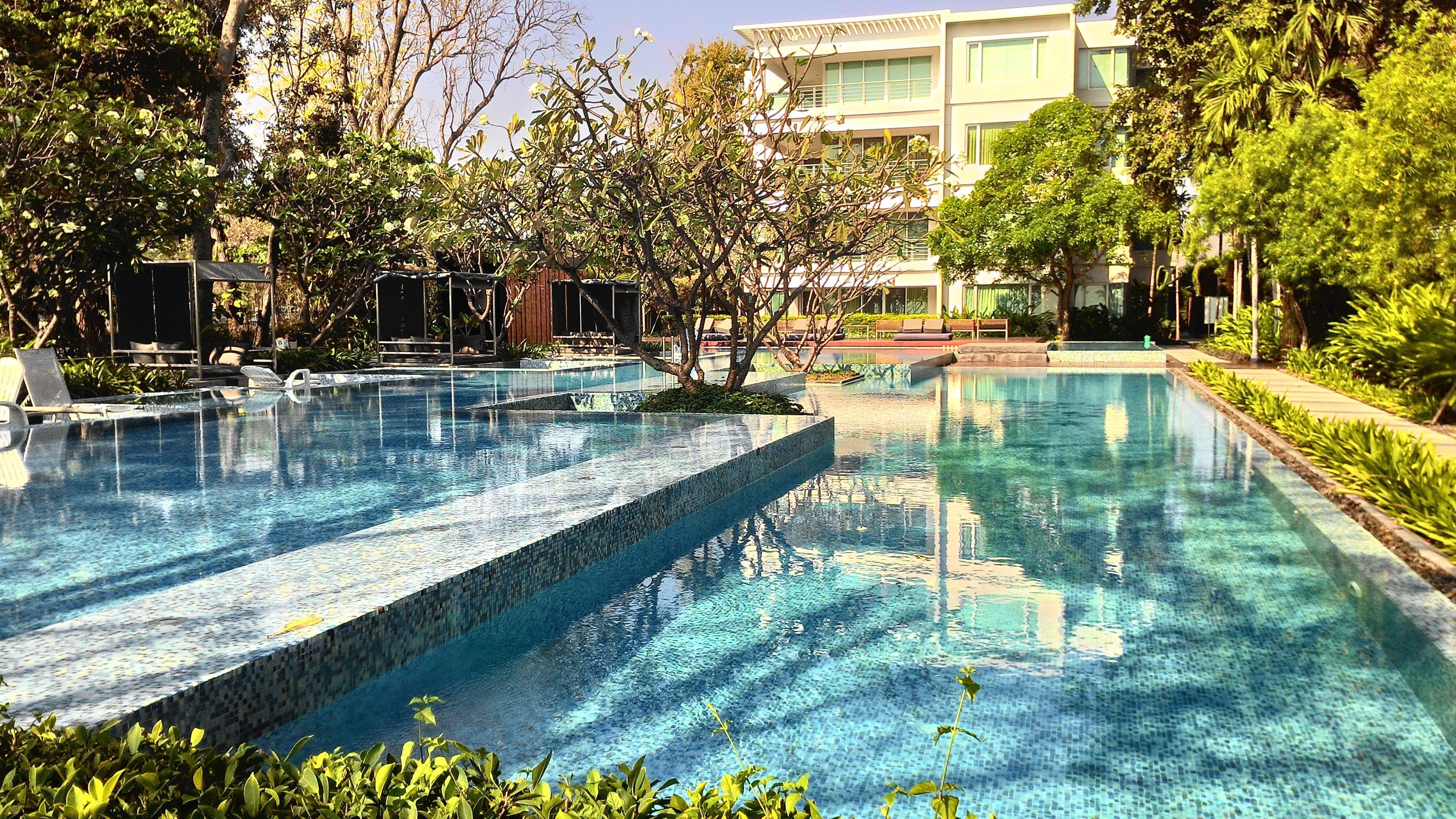A swimming pool with a tiled edge, surrounded by greenery and a building in the background.