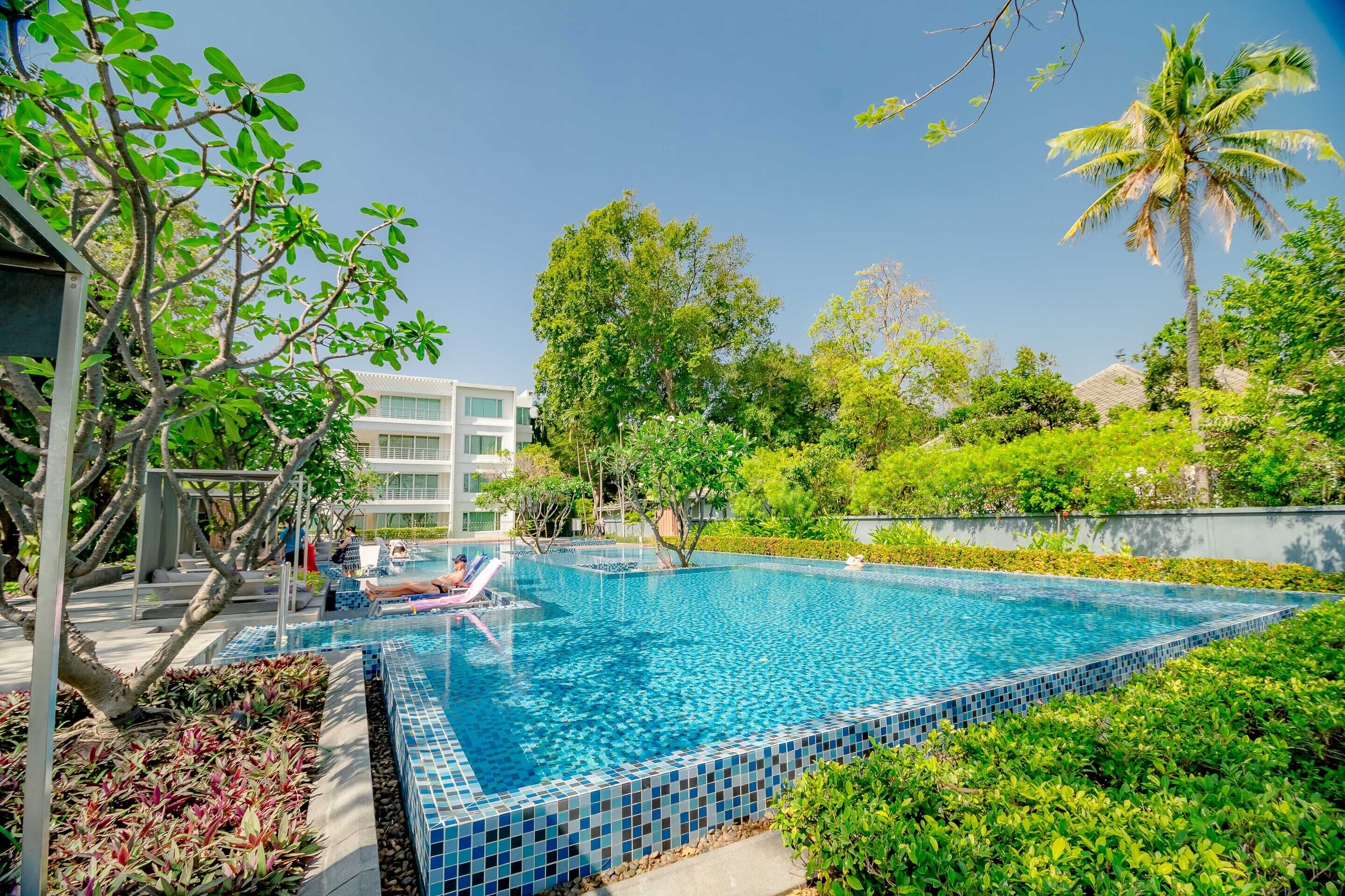 Una piscina con bordes de azulejos, rodeada de abundante vegetación y un edificio al fondo.