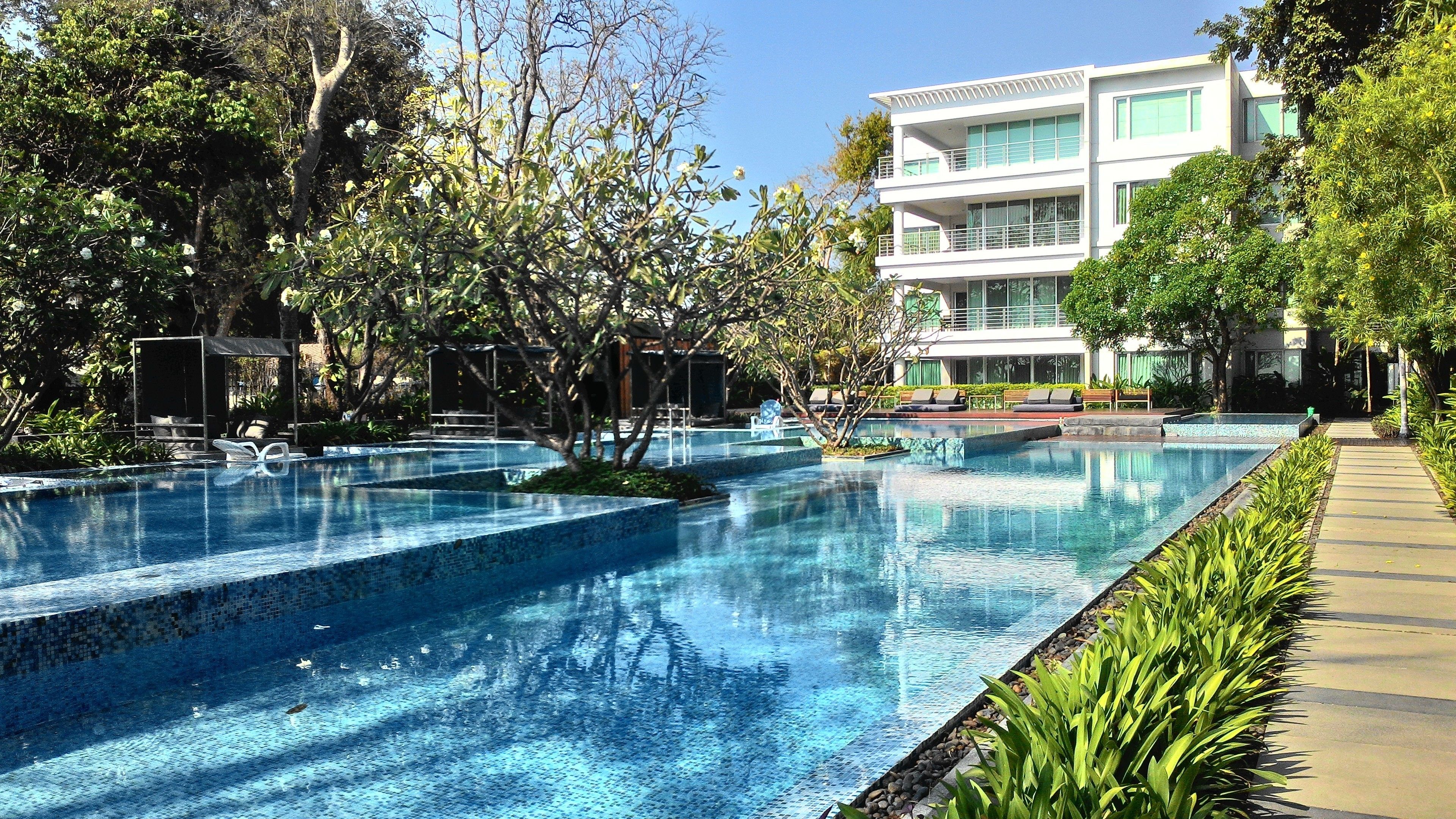 Una piscina con agua azul cristalina, rodeada de abundante vegetación y un edificio moderno con balcones.