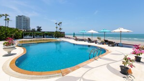 Ein Schwimmbecken mit gewölbter Kante, umgeben von Liegestühlen und Sonnenschirmen, mit Strand und Meerblick im Hintergrund.