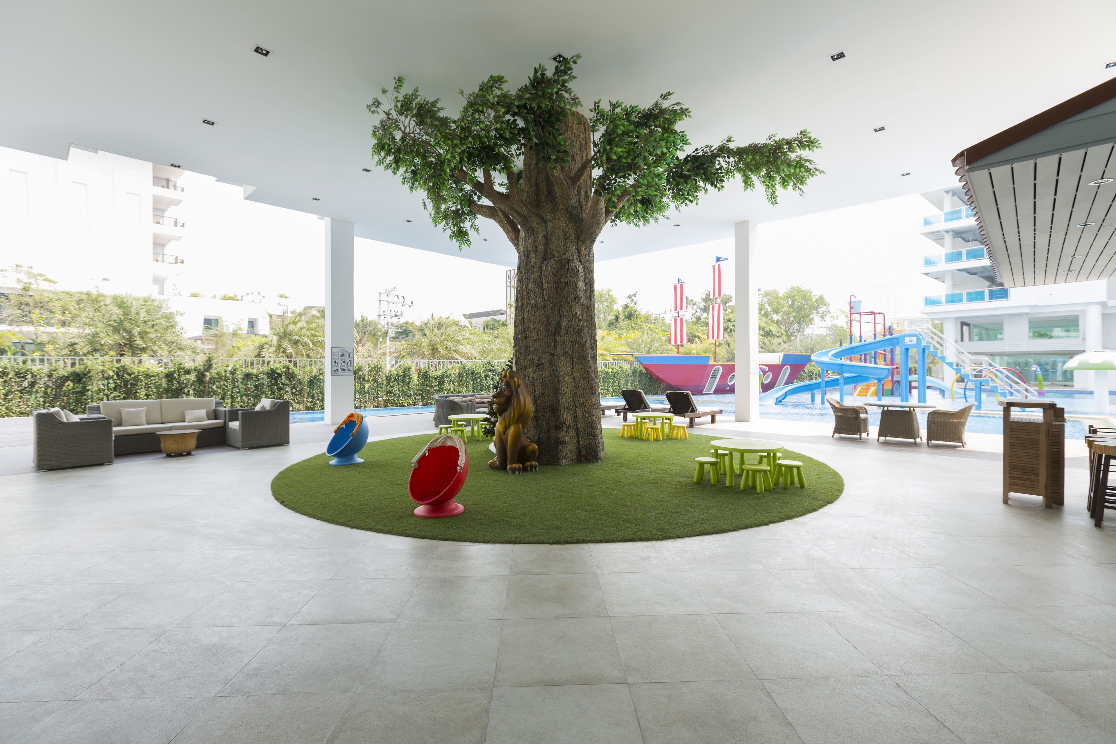 A spacious indoor area with a large artificial tree, seating arrangements, and a playground in the background.