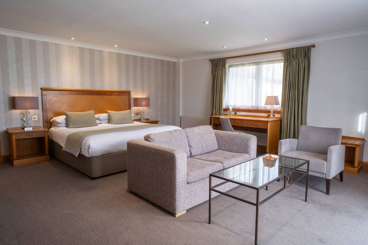 A hotel room with a bed, a sofa, a chair, a glass coffee table, a desk, and a lamp.