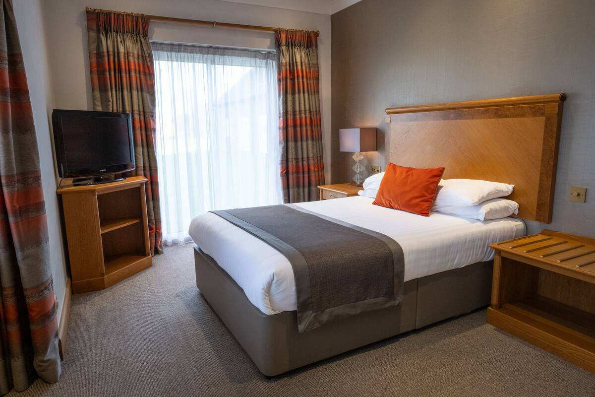 A hotel room with a large bed, a television on a wooden stand, a bedside table with a lamp, and curtains.