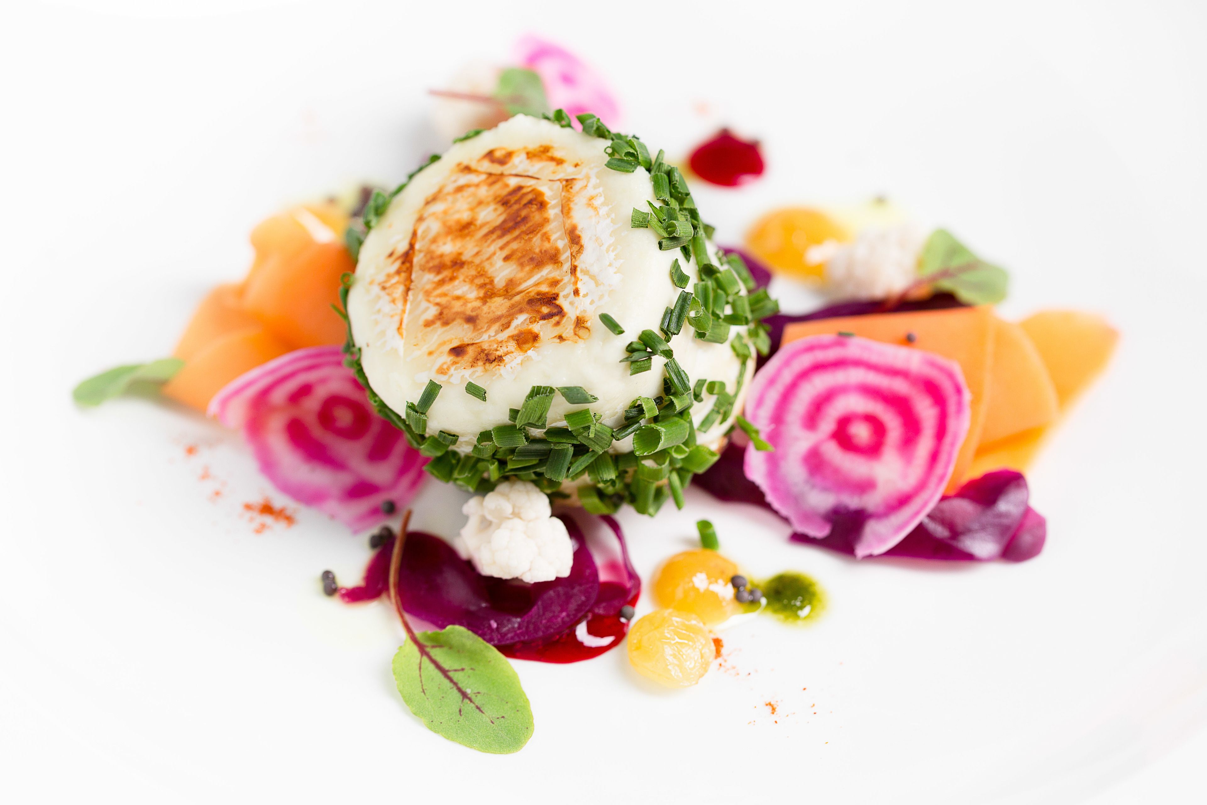 A white plate with a food dish consisting of a spherical item, possibly a cheese or a dumpling, garnished with green herbs, surrounded by slices of beetroot and other vegetables.