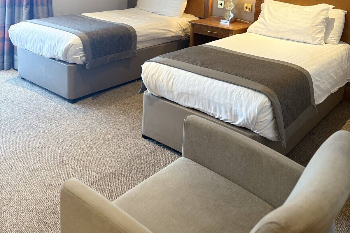 A hotel room with two beds, a nightstand with a lamp, a chair, and a window with curtains.
