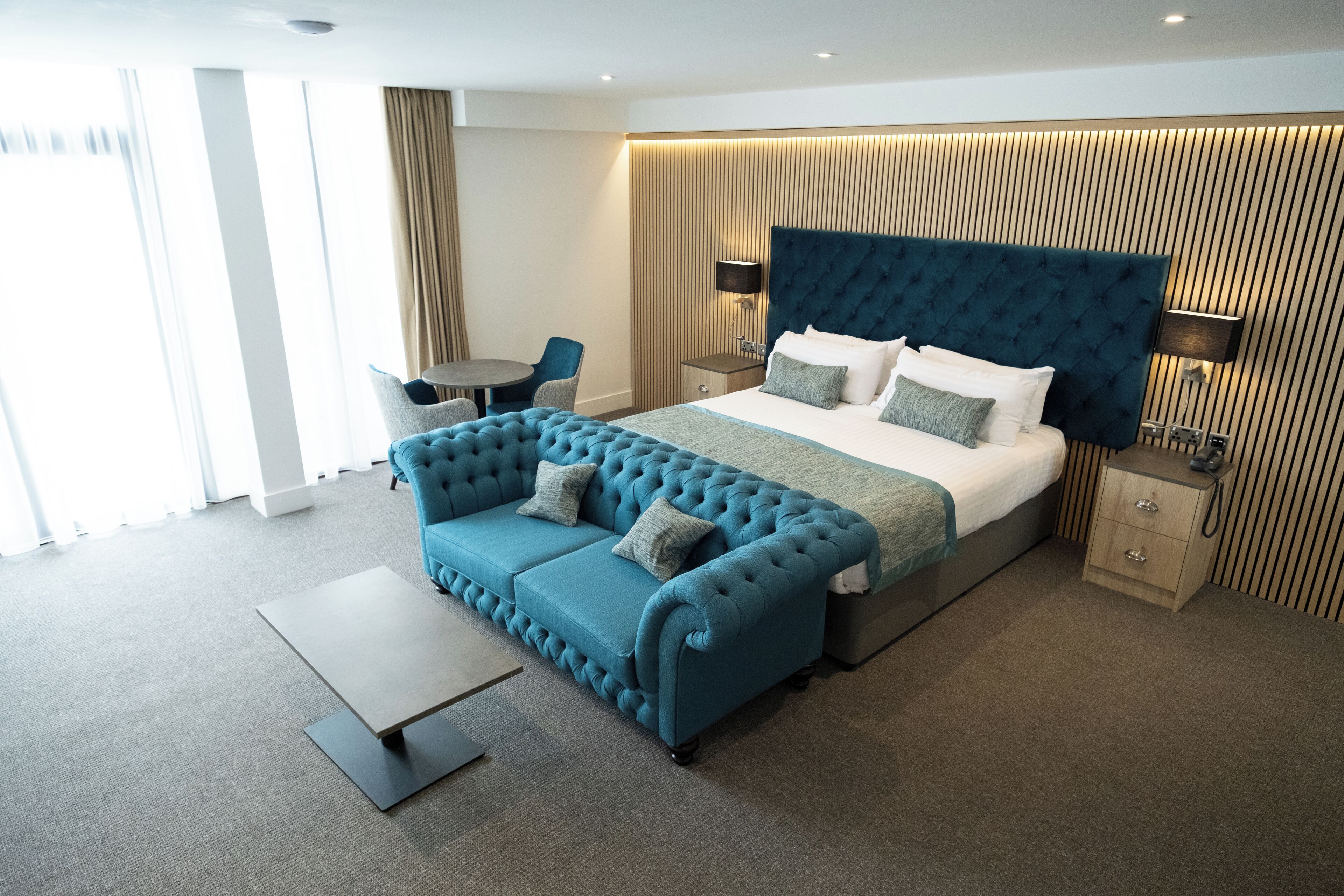 Ein modernes Hotelzimmer mit einem großen Bett, einem geblümten blauen Sofa, einem kleinen runden Tisch und einem Nachttisch mit einer Lampe.