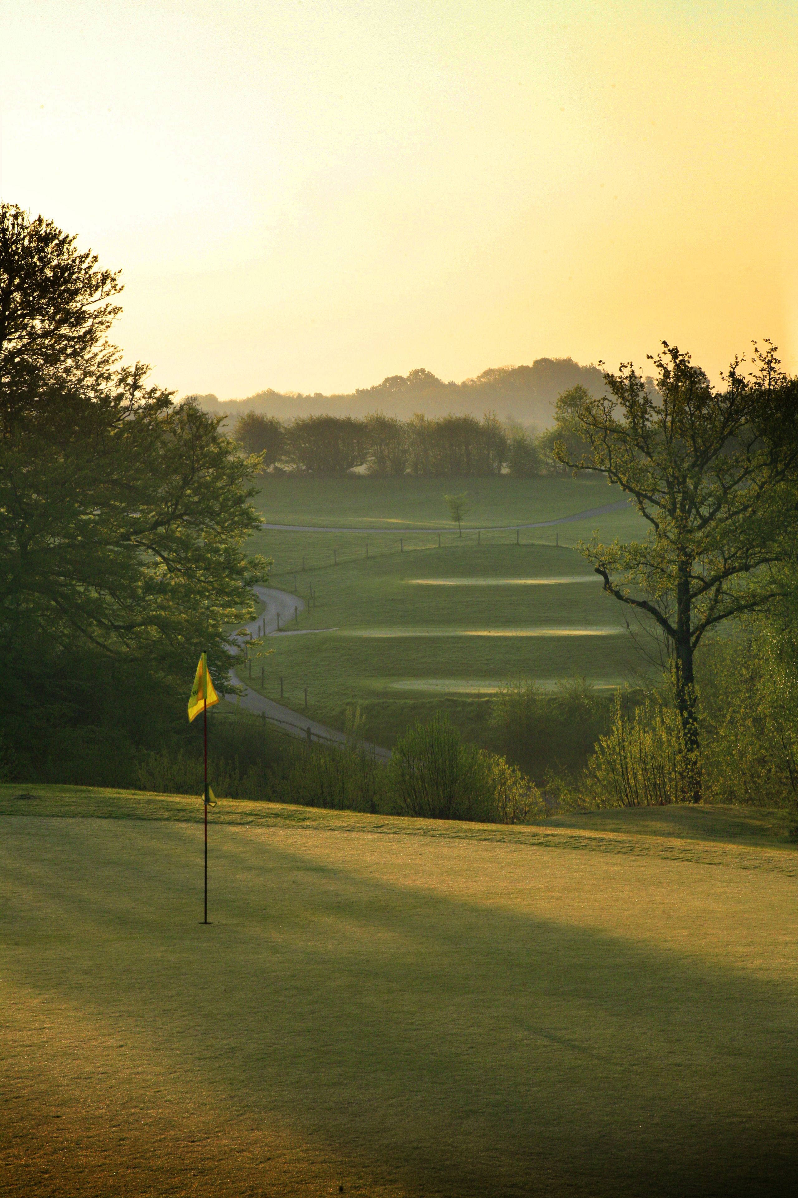A golf course with a flagstick in the foreground, surrounded by trees and rolling hills in the background.