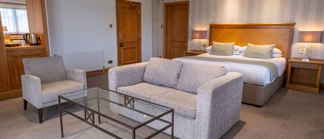 Habitación de hotel con cama, dos sofás, una mesa de centro de vidrio, puertas de madera y una zona de kitchenette.