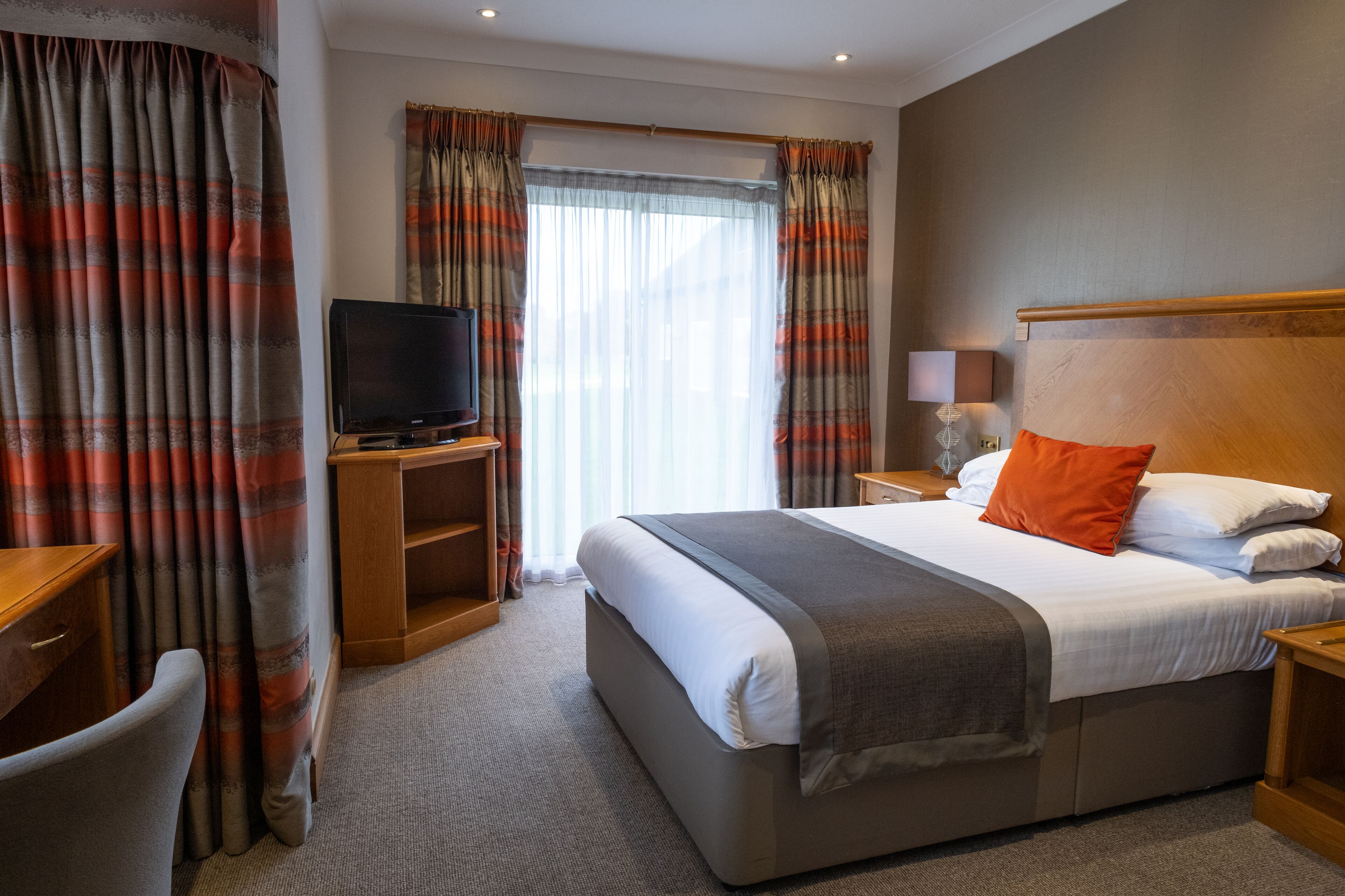 A hotel room with a large bed, a TV on a wooden stand, a desk with a chair, and a window with curtains.