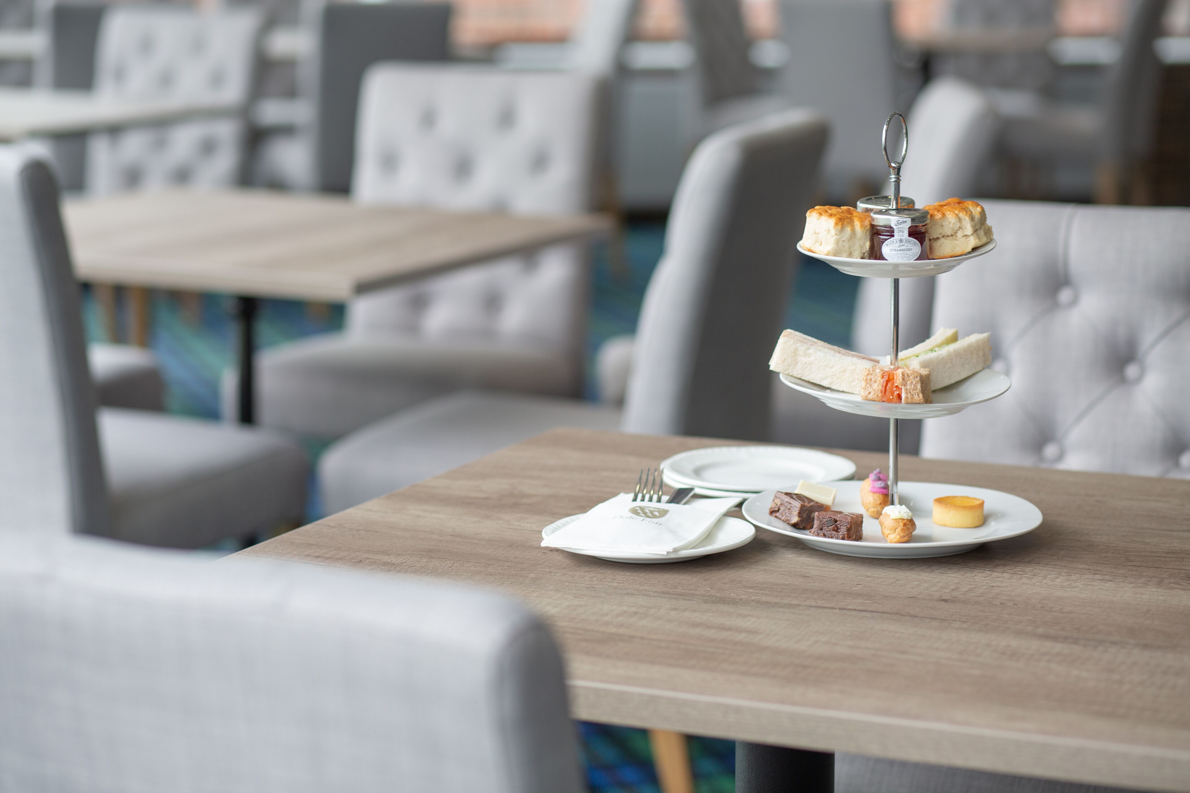 Un présentoir à trois niveaux, garni de sandwichs et de pâtisseries, est placé sur une table dans un coin repas, où se trouvent des chaises et des tables recouvertes de tissu.