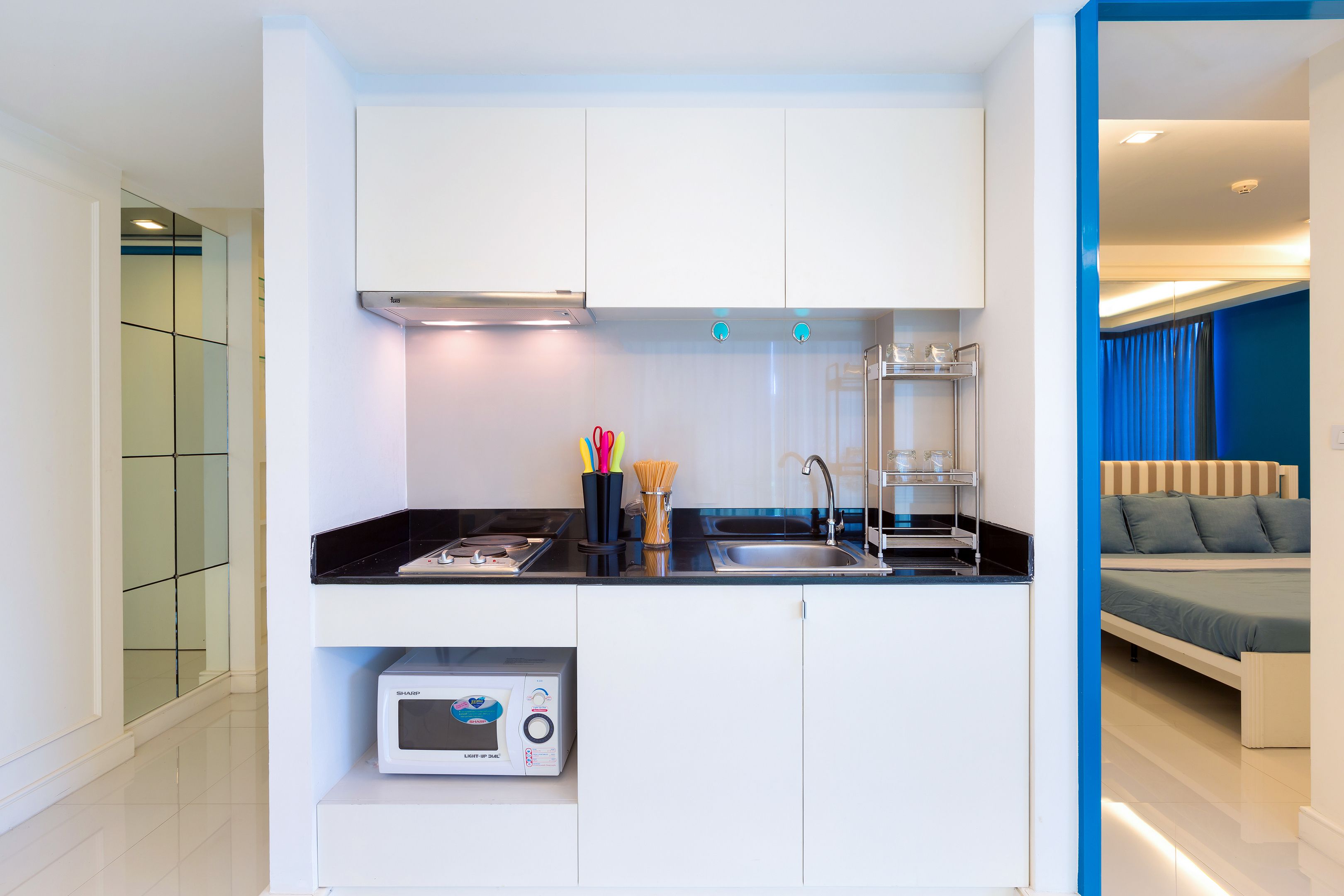 A modern kitchen with white cabinets, a black countertop, and a microwave. There is a sink with a faucet, and a bed with a blue curtain in the adjacent room.